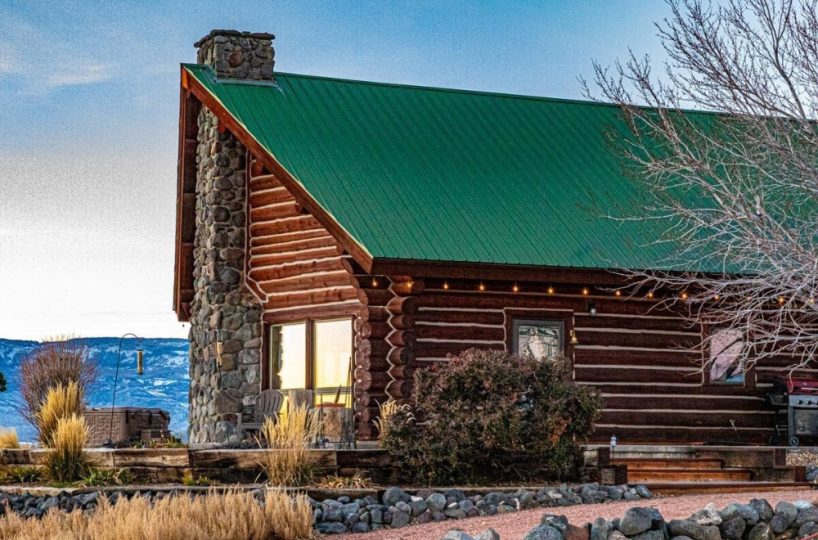 Rustic log home with views of Colorado mountains featuring stone chimney, green metal roof, string lights, and natural landscaping.