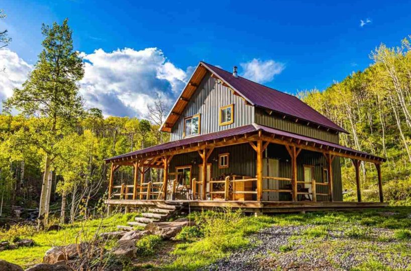 Rustic cabin in the woods with wrap-around porch, surrounded by aspen trees and vibrant nature, under blue sky with fluffy clouds.