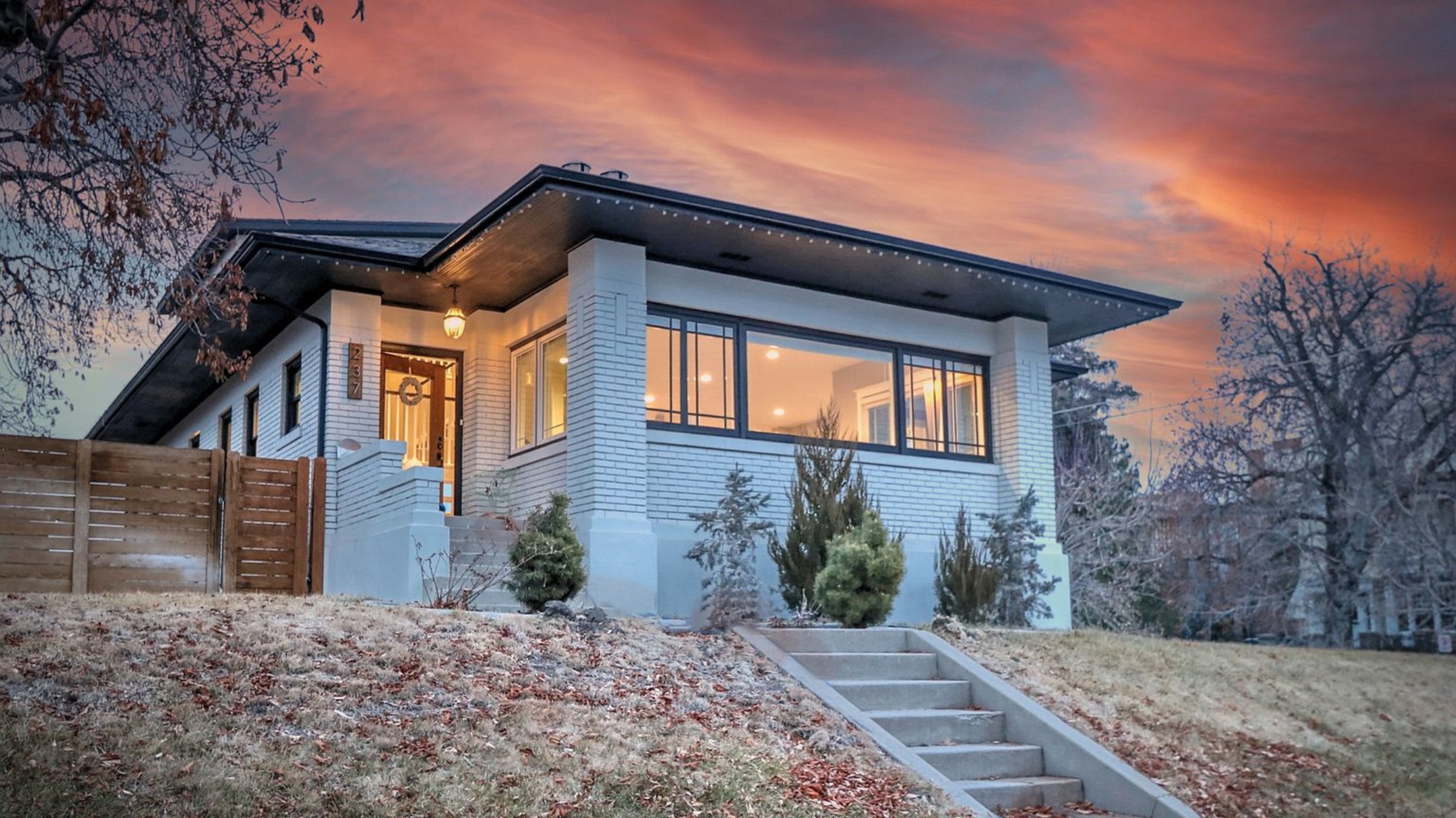 Modern white Prairie-style home with large windows and horizontal lines, illuminated at sunset with orange and pink sky. Features clean architectural details, horizontal wood privacy fence, concrete steps, and contemporary landscape design. Property number 234 visible.
